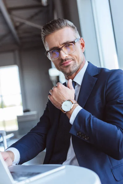 Bel homme d'affaires dans des lunettes en utilisant un ordinateur portable et montrant montre à l'aéroport — Photo de stock