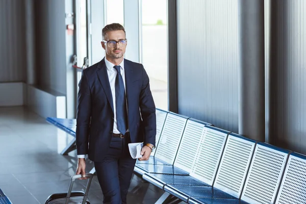 Adulto guapo hombre de negocios en gafas con equipaje y billetes caminando en el aeropuerto - foto de stock