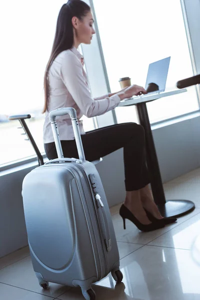 Bolsa de equipaje gris y joven empresaria escribiendo en el portátil en el aeropuerto - foto de stock