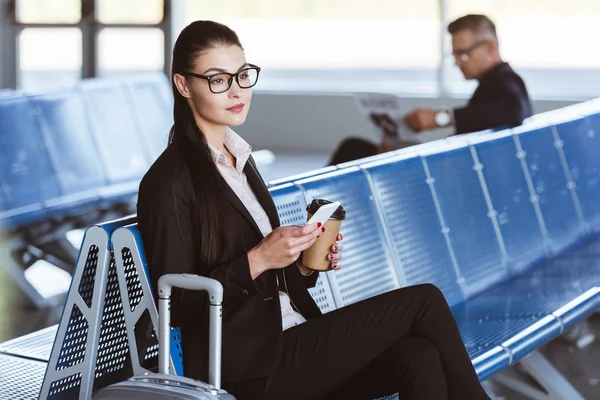 Молодая бизнесвумен в очках, сидящая со смартфоном и кофе в аэропорту — стоковое фото