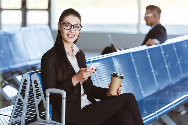 Joven mujer de negocios sonriente en vasos con café para ir con teléfono inteligente en la sala de salida en el aeropuerto - foto de stock
