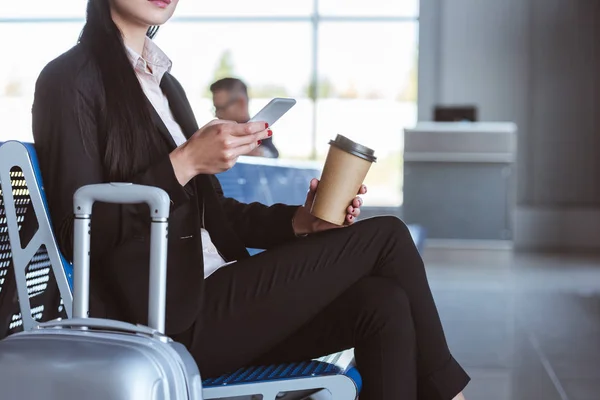 Close up of young businesswoman with coffee to go using smartphone in departure lounge at airport — Stock Photo
