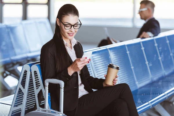 Jovem mulher de negócios sorridente em óculos com café para ir usando smartphone na sala de embarque no aeroporto — Fotografia de Stock