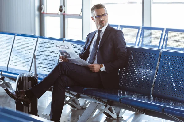 Hombre de negocios adulto con gafas sentado en la sala de salida en el aeropuerto y leyendo el periódico - foto de stock