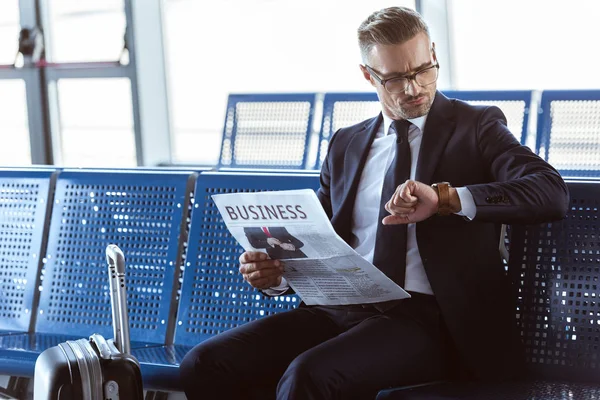 Erwachsener Geschäftsmann sitzt mit Zeitung in der Abflughalle am Flughafen und schaut auf die Uhr — Stockfoto