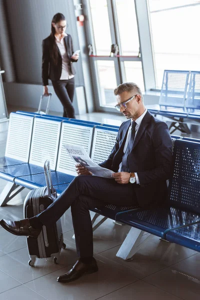 Adult handsome businessman reading newspaper while businesswoman walking at departure lounge at airport — Stock Photo