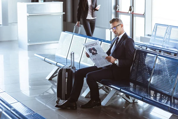 Adulto hombre de negocios leyendo periódico mientras que la mujer de negocios caminando en la sala de salida en el aeropuerto - foto de stock