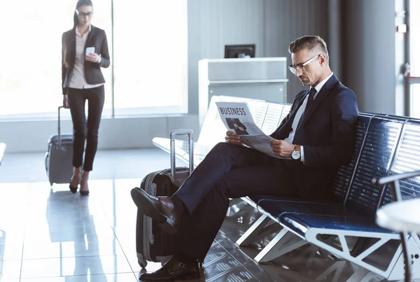 Adulto hombre de negocios leyendo el periódico mientras que la mujer de negocios caminando con el equipaje en la sala de salida en el aeropuerto - foto de stock