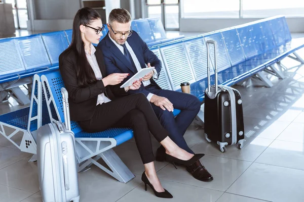 Adult businessman and businesswoman sitting at departure lounge in airport and using digital tablet — Stock Photo