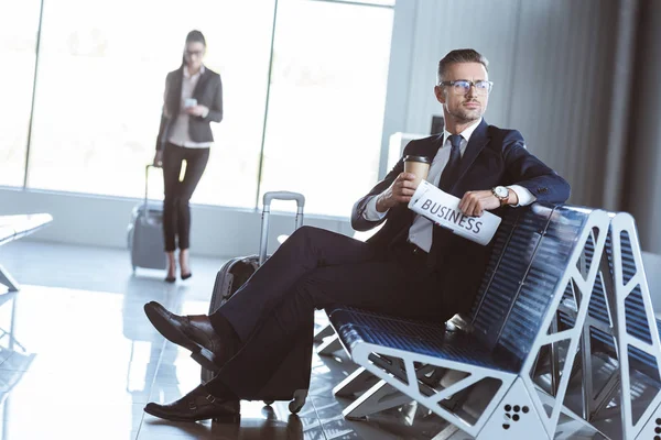 Erwachsener Geschäftsmann sitzt in Abflughalle, während Geschäftsfrau auf Flughafen geht — Stockfoto