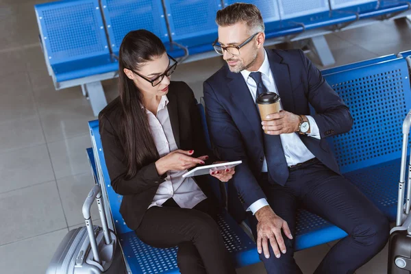 Erwachsener Geschäftsmann trinkt Kaffee, während Geschäftsfrau digitales Tablet in der Abflughalle am Flughafen benutzt — Stockfoto