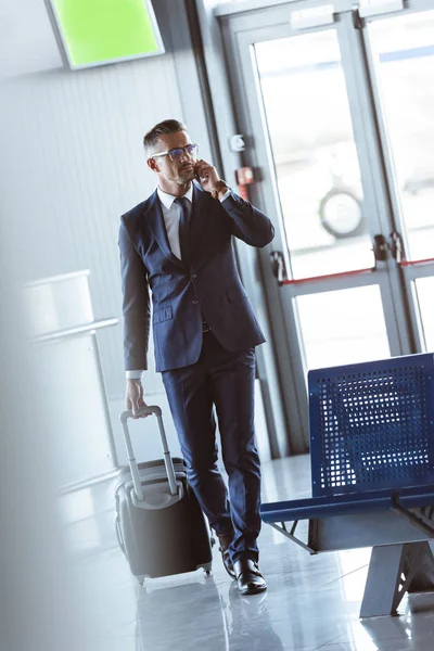Adulto guapo hombre de negocios con teléfono inteligente y equipaje caminando en el aeropuerto - foto de stock
