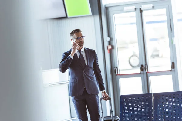 Erwachsene gut aussehende Geschäftsmann mit Smartphone und Gepäck zu Fuß am Flughafen — Stockfoto