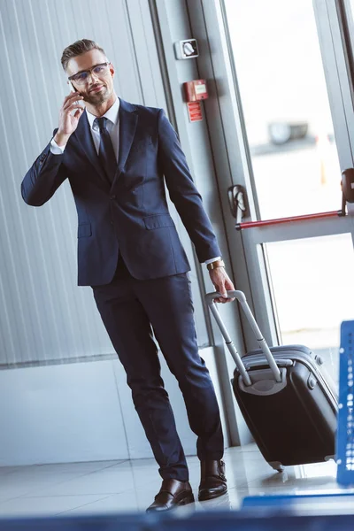 Adulto guapo hombre de negocios en gafas caminando en el aeropuerto y hablando en smartphone - foto de stock