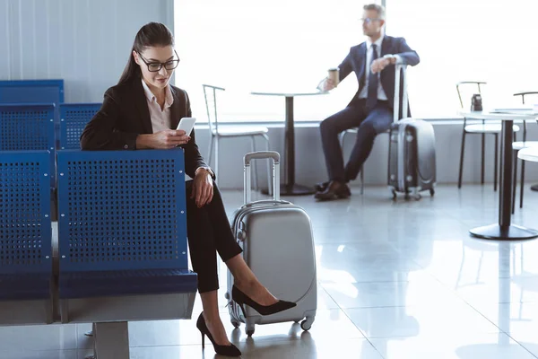 Junge Geschäftsfrau nutzt Smartphone, während Geschäftsmann mit Coffee to go im Flughafen sitzt — Stockfoto