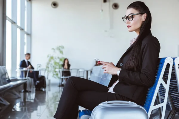 Jeune femme d'affaires en lunettes en utilisant smartphone au salon de départ à l'aéroport — Photo de stock
