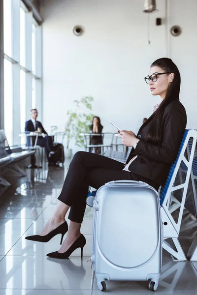 Joven mujer de negocios en gafas con teléfono inteligente sentado en la sala de salida en el aeropuerto - foto de stock
