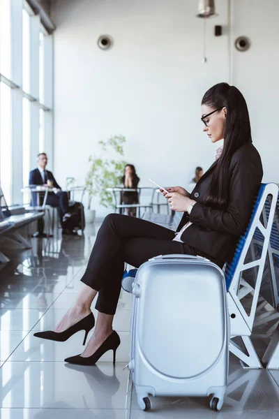 Молодая деловая женщина в очках со смартфоном сидит в зале вылета в аэропорту — стоковое фото