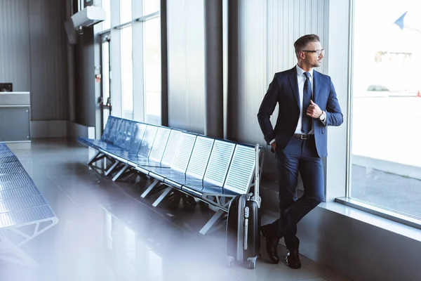 Hombre de negocios guapo de pie en la sala de salida y mirando a la ventana en el aeropuerto - foto de stock