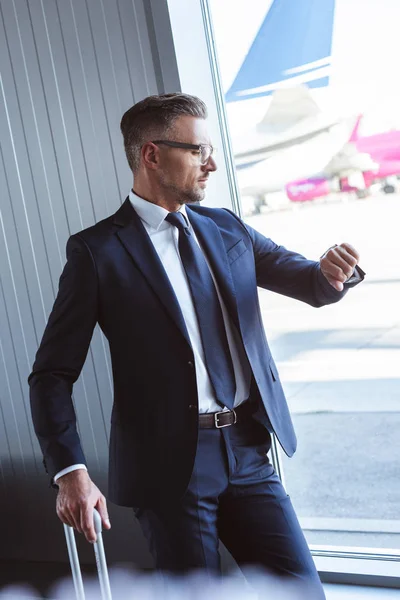 Uomo d'affari in occhiali in piedi presso la sala di partenza vicino alla finestra e guardando orologio in aeroporto — Foto stock
