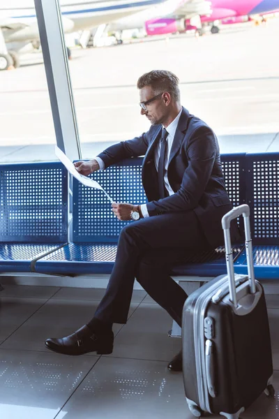 Bel homme d'affaires dans des lunettes assis près de la fenêtre au salon de départ et le journal de lecture — Photo de stock