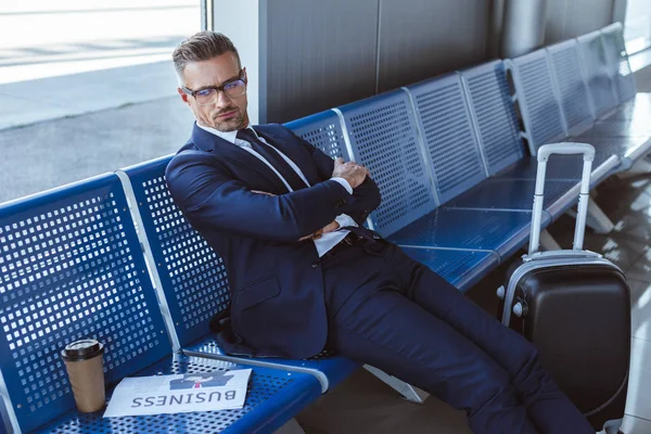 Hombre de negocios cansado en gafas sentado cerca de la ventana con periódico y taza de papel en el aeropuerto - foto de stock