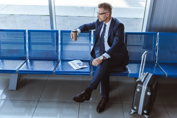 Hombre de negocios en vasos sentado cerca de la ventana con periódico y café para ir en el aeropuerto - foto de stock