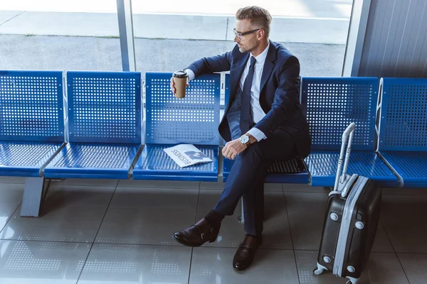 Uomo d'affari in bicchieri seduto vicino alla finestra con giornale e caffè per andare in aeroporto — Foto stock