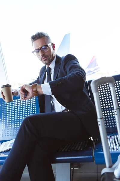 Empresário em óculos sentado perto da janela com café para ir e olhando para assistir no aeroporto — Fotografia de Stock