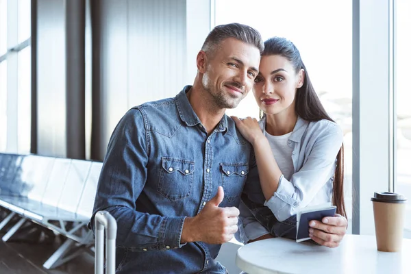 Beautiful couple sitting in the airport with coffee and man showing thumb up — Stock Photo