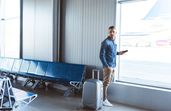 Hombre con maleta de plata revisando el teléfono y esperando cerca de la ventana en el aeropuerto - foto de stock