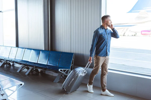 Hombre guapo con equipaje plateado hablando por teléfono y caminando en el aeropuerto - foto de stock