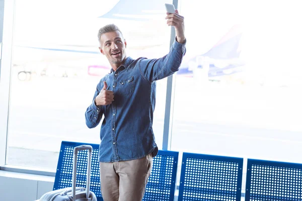 Человек, сидящий в аэропорту и улыбающийся, показывающий большой палец и делающий селфи на смартфоне — стоковое фото