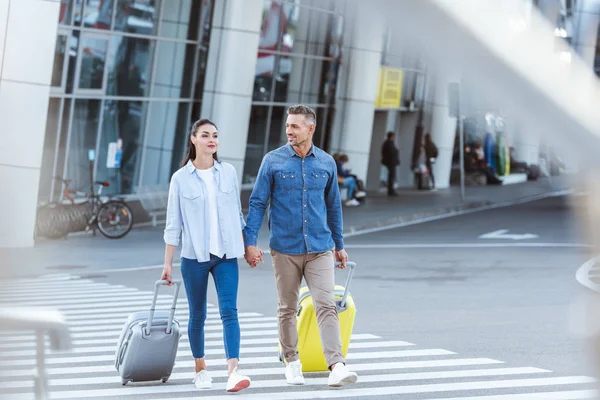 A couple of tourists crossing pedestrian, holding hands and pulling their luggage — Stock Photo