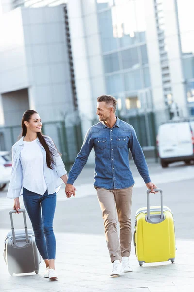 Un par de turistas tomados de la mano, tirando de su equipaje, mirándose unos a otros y sonriendo en el fondo del aeropuerto - foto de stock