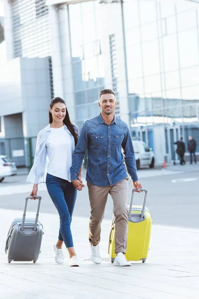 Um casal de viajantes atravessando pedestres, de mãos dadas e puxando sua bagagem — Fotografia de Stock