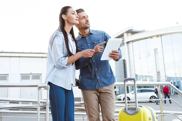 Couple of tourists with luggage holding digital tablet, pointing and looking away — Stock Photo