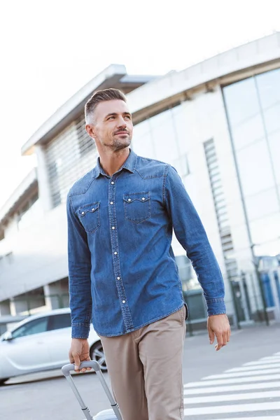 Guapo sonriente hombre adulto cruzando peatón y tirando de su equipaje - foto de stock