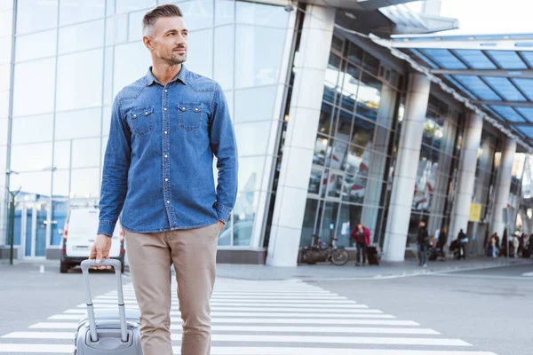 Schöner erwachsener Mann überquert Fußgänger, zieht sein Gepäck, schaut weg und lächelt auf dem Hintergrund des Flughafens — Stockfoto