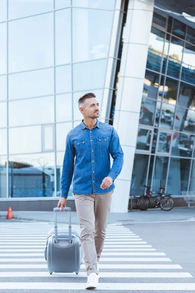 Handsome smiling man crossing pedestrian, pulling his luggage and looking away — Stock Photo