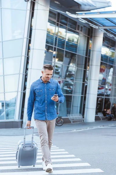 Hombre adulto guapo con equipaje cruzando el peatón, mirando en el teléfono inteligente en el fondo del aeropuerto - foto de stock