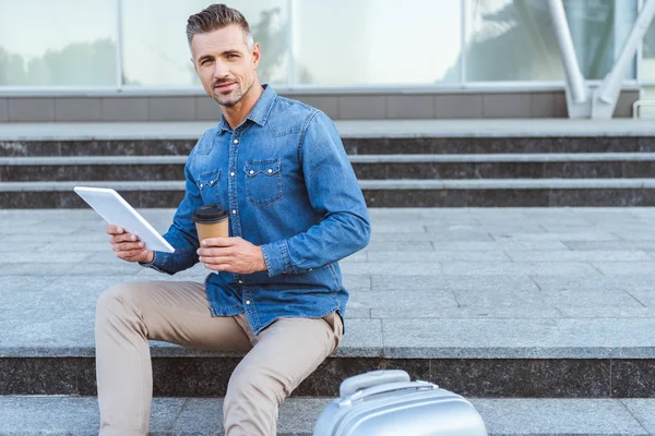 Schöner erwachsener Mann mit Kaffeetasse, der auf der Treppe sitzt, ein digitales Tablet in der Hand hält und in die Kamera lächelt — Stockfoto