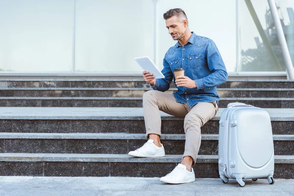 Bello uomo adulto con un caffè per andare seduto sulle scale con i bagagli e utilizzando tablet digitale — Foto stock