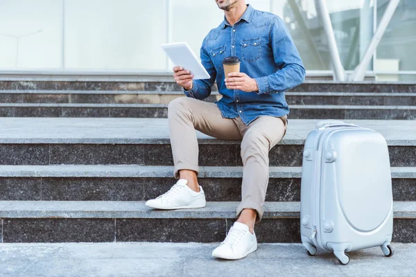 Man sitting on the staircase with luggage, holding digital tablet and coffee to go — Stock Photo