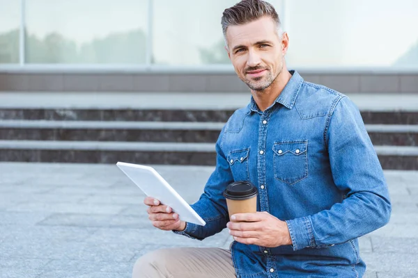 Hombre adulto guapo con una taza de café sosteniendo la tableta digital, mirando a la cámara y sentado en la escalera - foto de stock