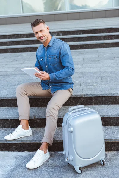 Handsome adult man using digital tablet and smiling at camera while sitting on the staircase with luggage — Stock Photo