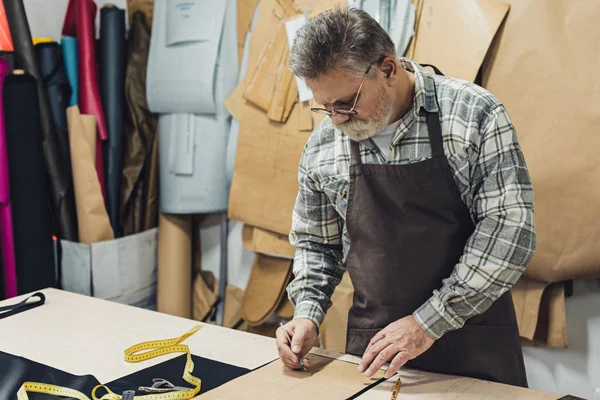 Mature male leather handbag craftsman in apron and eyeglasses working at studio — Stock Photo