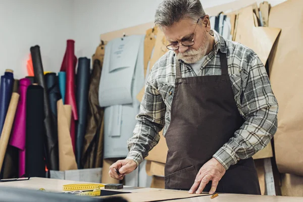 Selective focus of mature leather handbag craftsman in apron and eyeglasses working at studio — Stock Photo