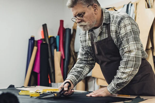 Sac à main concentré artisan en tablier et lunettes découpe cuir par ciseaux à l'atelier — Photo de stock