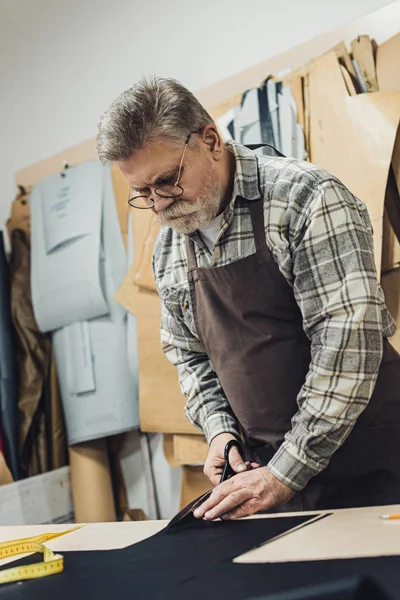 Mature handbag craftsman in apron and eyeglasses cutting leather by scissors at workshop — Stock Photo